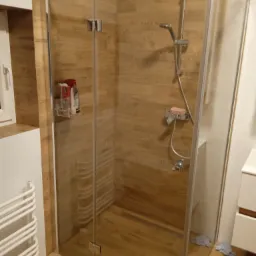 Kabina prysznicowa ze szklanymi drzwiami, wykończona płytkami imitującymi drewno, z widoczną armaturą i półką na kosmetyki, uzupełniona o grzejnik drabinkowy i szare ręczniki na podłodze.