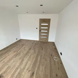 Jasne panele podłogowe ułożone w pustym pokoju z białymi ścianami, widoczne gniazdka elektryczne i drewniane drzwi z przeszkleniami.