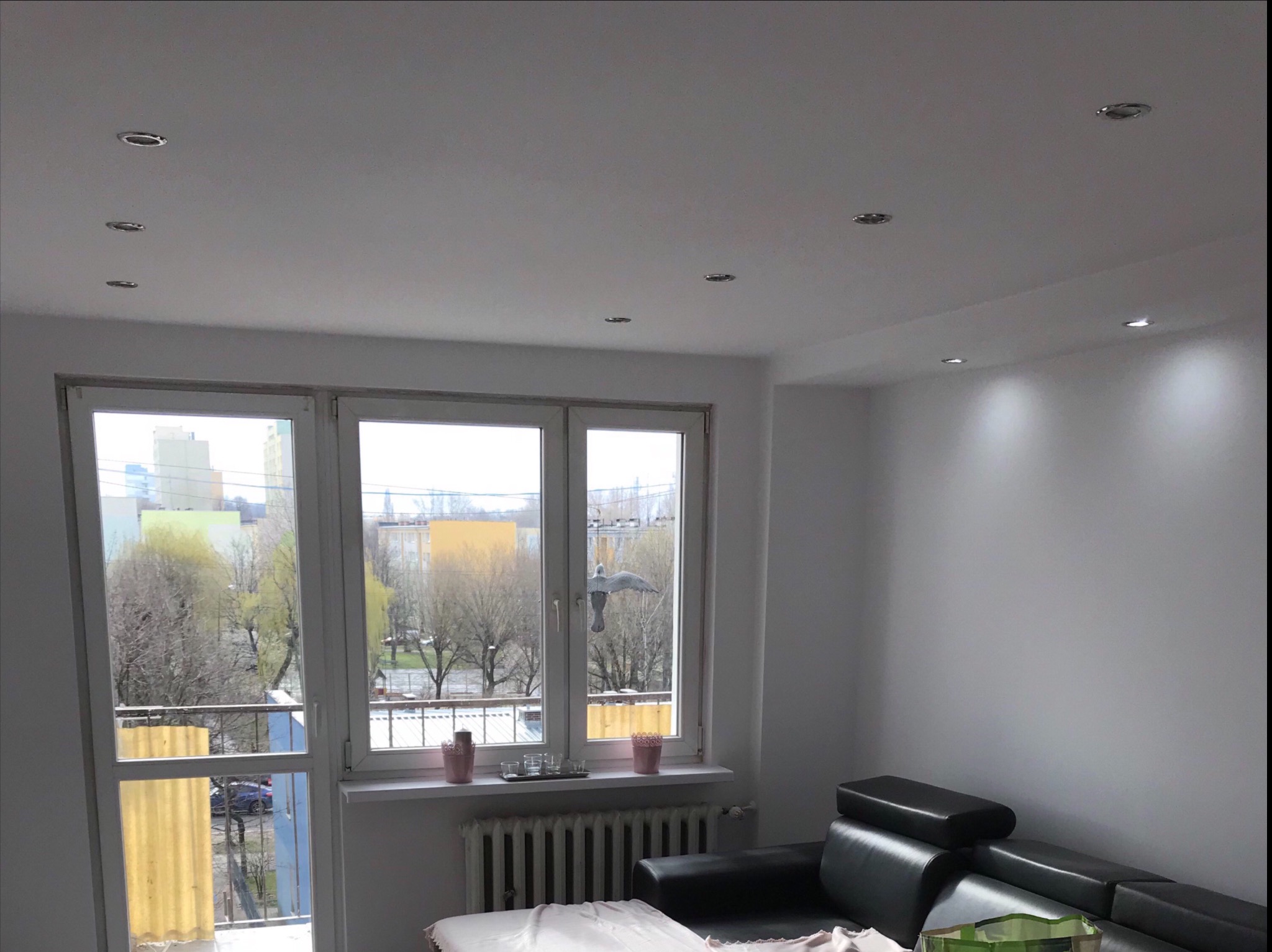 Salon po odświeżeniu: białe ściany, sufit z punktowym oświetleniem, okno z widokiem na drzewa i balkon, czarna skórzana sofa w rogu.