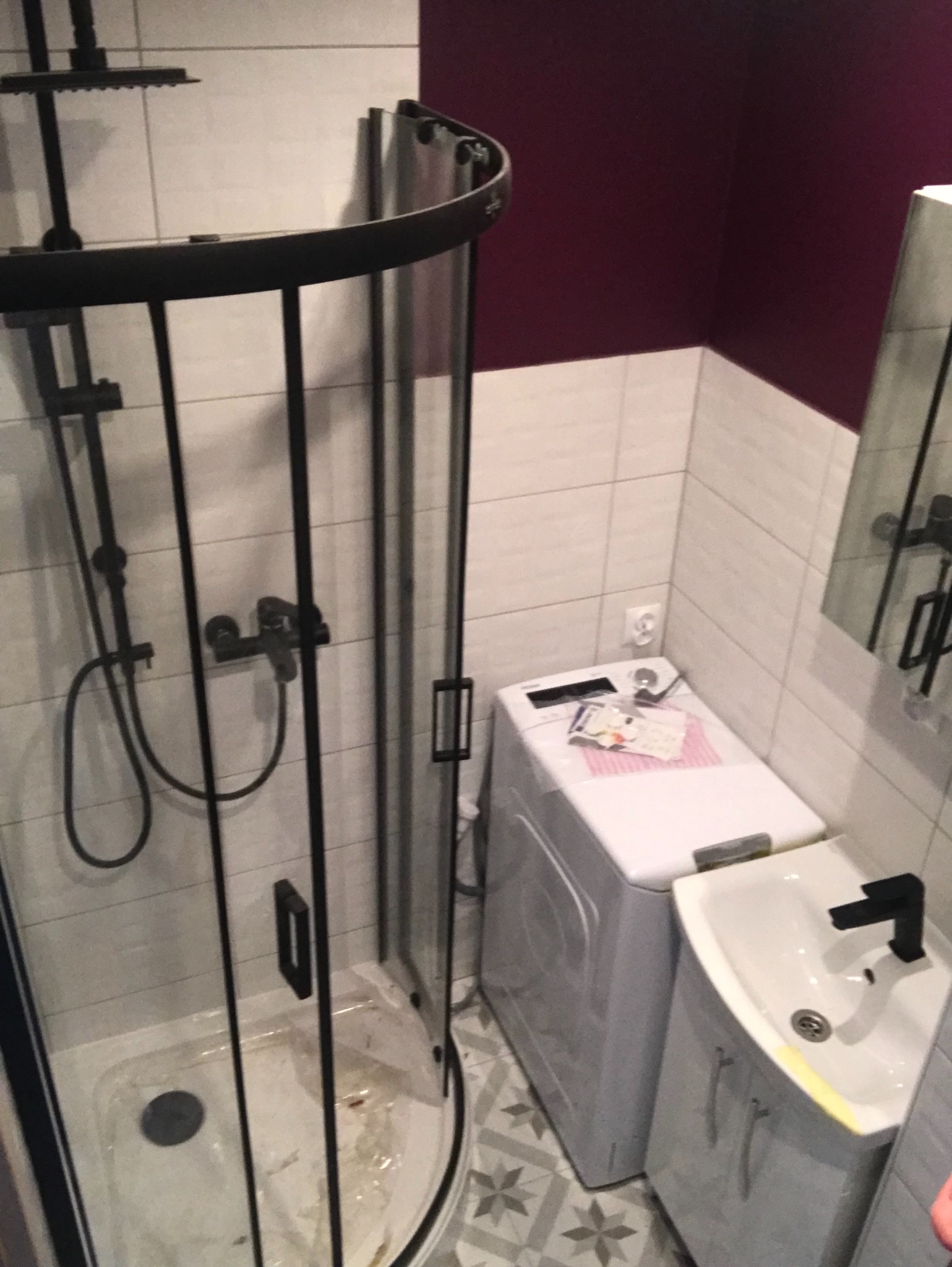 Ciasna łazienka z kabiną prysznicową z czarnymi profilami, białą umywalką narożną z czarną armaturą i pralką. Na pralce leżą dokumenty lub rachunki.