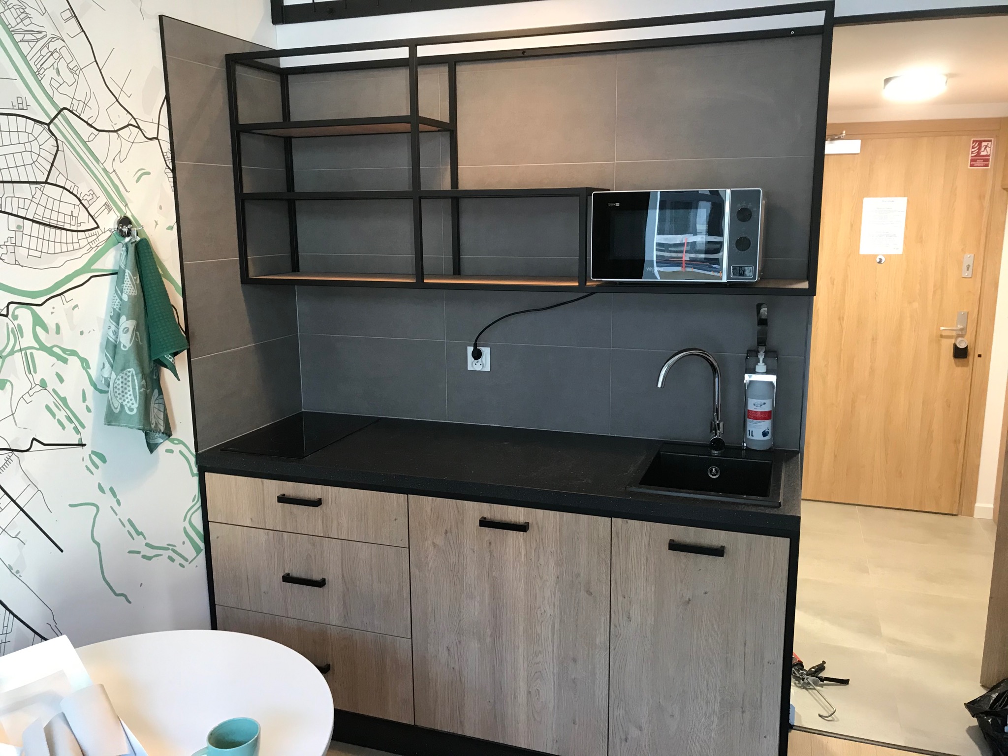 Nowoczesna, kompaktowa kuchnia z drewnianymi szafkami, czarnym blatem, mikrofalówką i otwartymi półkami z metalową ramą; na ścianie tapeta z mapą, obok biały okrągły stół i turkusowy kubek.