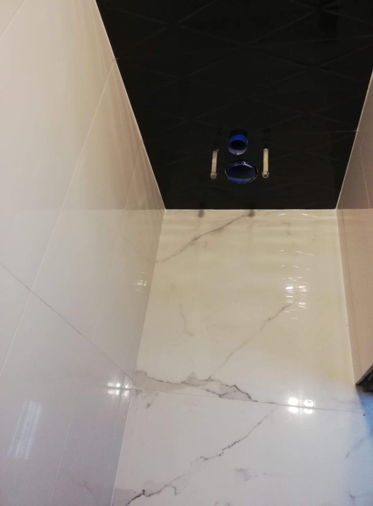 Białe, błyszczące płytki imitujące marmur na ścianach i podłodze, czarny sufit z geometrycznym wzorem, widoczne rury instalacji sanitarnej w trakcie montażu.