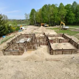 Szeroki kadr na wykop pod fundamenty domu jednorodzinnego z drewnianym szalunkiem, widoczny częściowo wylany beton w jednym z wykopów, w tle drzewa, koparka i inne maszyny budowlane.