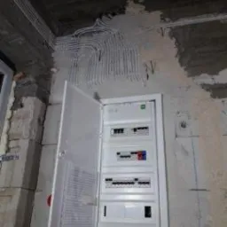 Otwarta skrzynka elektryczna z bezpiecznikami na surowej ścianie z widocznymi przewodami elektrycznymi biegnącymi po suficie, w trakcie montażu.