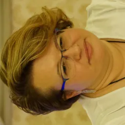 Portret kobiety w okularach, w białej bluzce, z blond włosami i srebrnymi kolczykami, na rozmytym tle w ciepłych odcieniach. Ujęcie z bliska, skupione na twarzy.