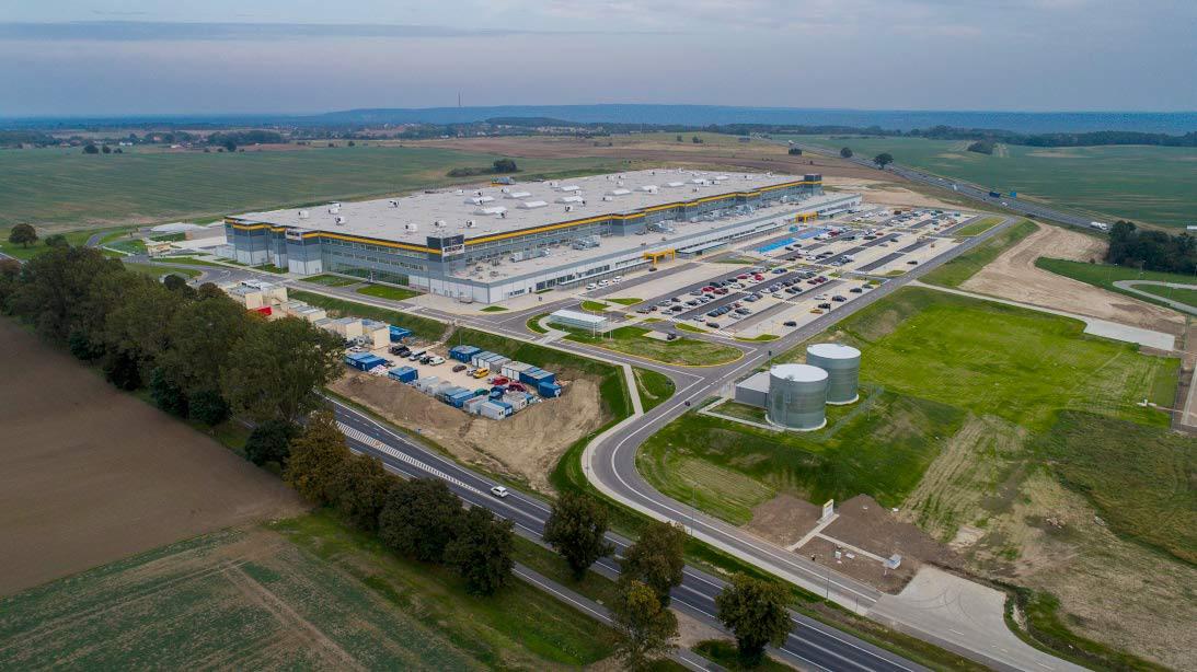 Szeroki kadr z lotu ptaka na rozległy, nowo wybudowany magazyn z dużym parkingiem, zbiornikami i drogami dojazdowymi, otoczony polami i lasami.