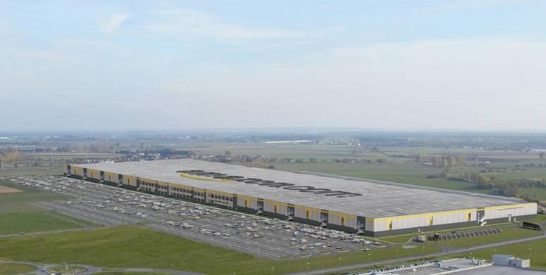 Rozległy magazyn z napisem na dachu, widok z lotu ptaka na tle krajobrazu.