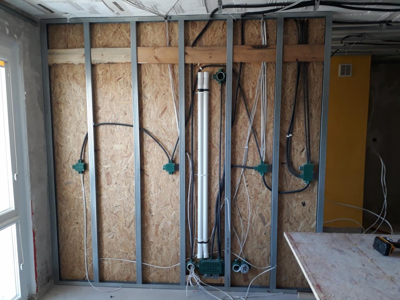 Instalacja elektryczna i wodno-kanalizacyjna w ścianie szkieletowej z płyt OSB, widoczne puszki instalacyjne, rury PCV i przewody elektryczne przed tynkowaniem.