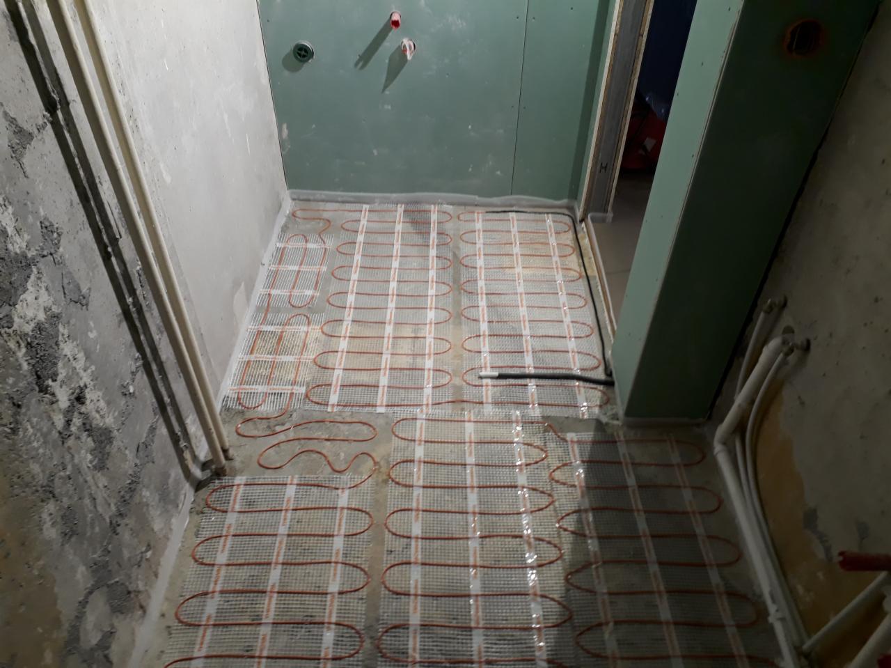 Rozłożona mata grzewcza na podłodze w surowym stanie, przygotowana do zalania wylewką, widoczne rury instalacyjne na ścianach.