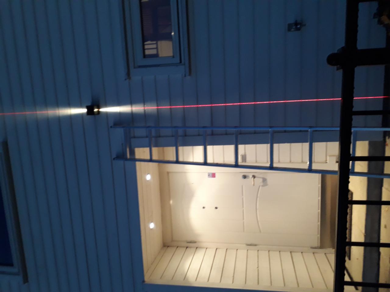 Montaż oświetlenia zewnętrznego: widoczna ściana budynku z zamontowaną lampą, z której biegnie czerwona linia lasera, oraz drabina oparta o ścianę, oświetlająca niszę z halogenami.