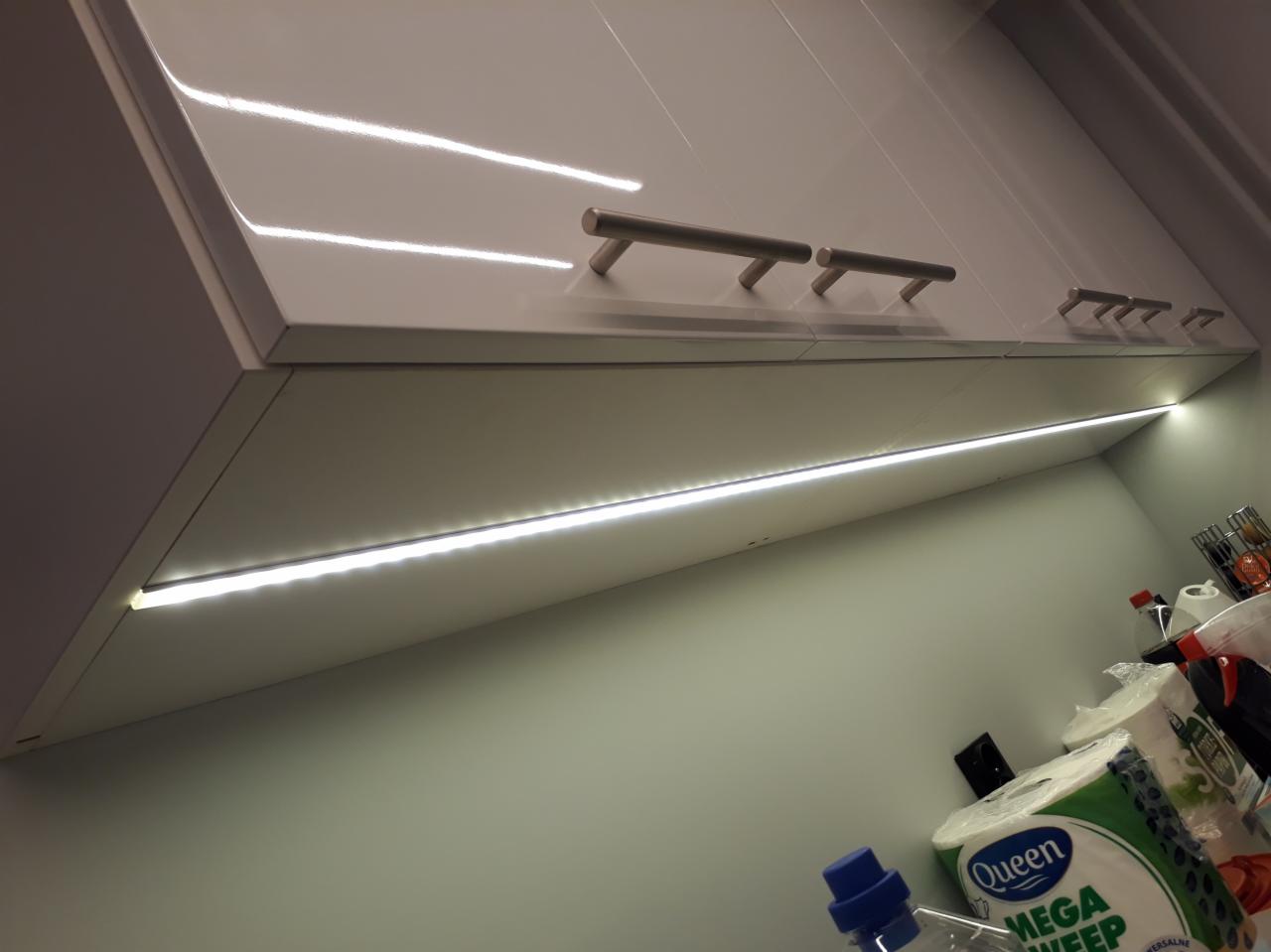 Białe, lakierowane szafki kuchenne z podświetleniem LED podszafkowym, widoczne srebrne uchwyty i fragment blatu z akcesoriami kuchennymi.