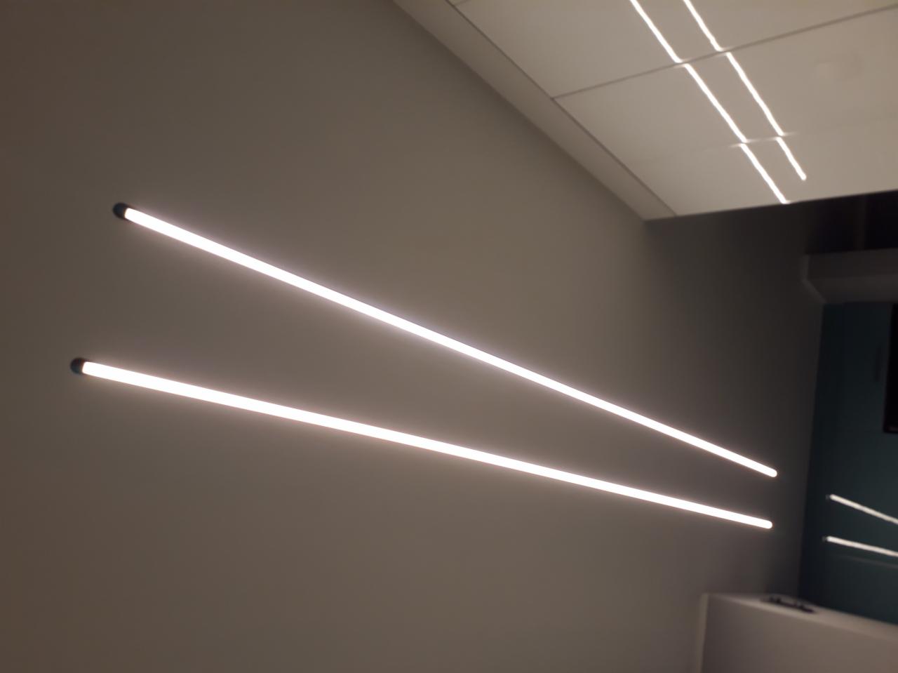 Dwie równoległe, podłużne lampy LED zamontowane poziomo na szarej ścianie, oświetlające wnętrze z widocznym fragmentem sufitu kasetonowego i elementami wyposażenia w tle.