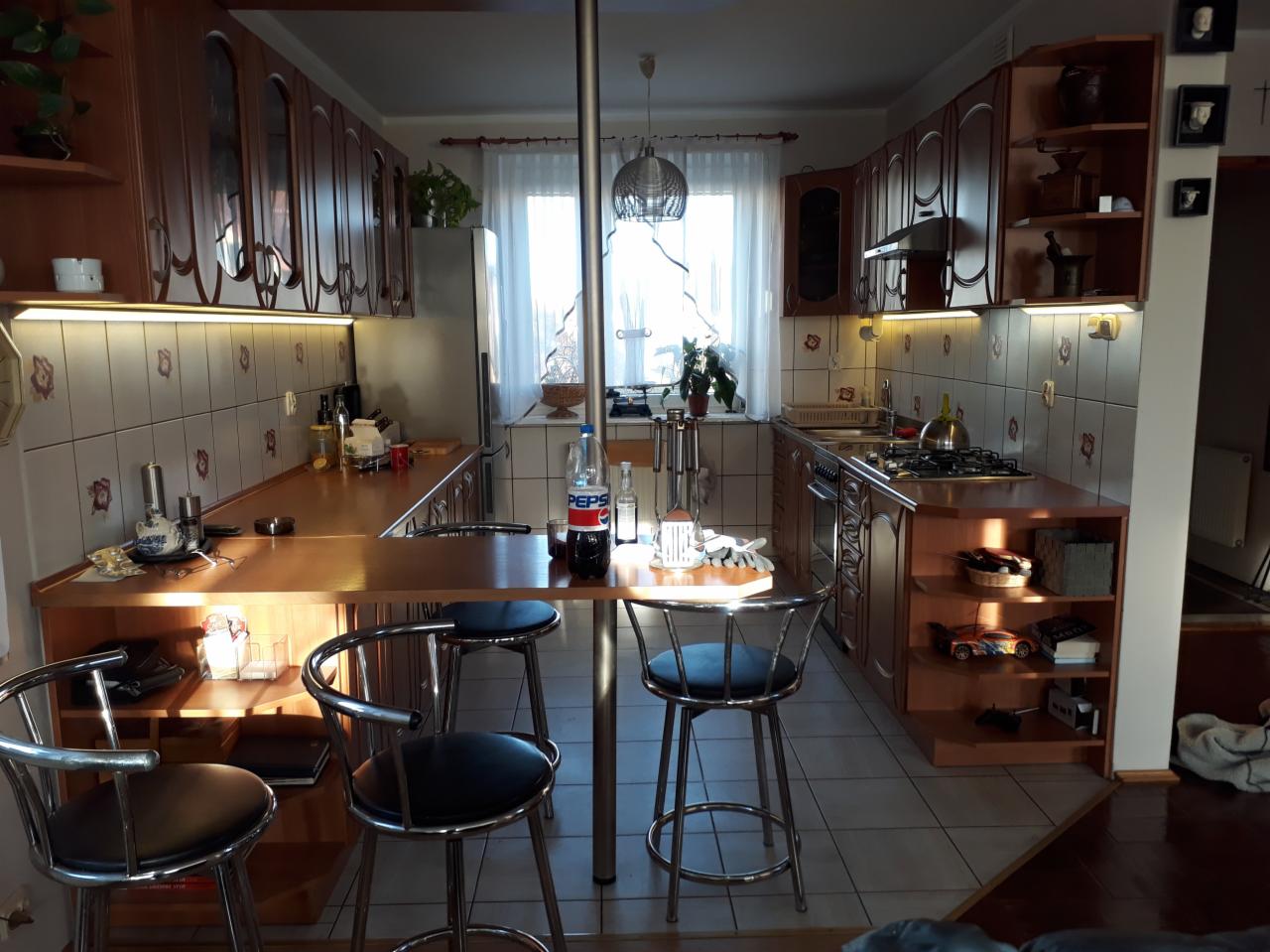 Ustawna kuchnia z drewnianymi szafkami i płytkami w kwiaty, oświetleniem podszafkowym i stołem z krzesłami barowymi na pierwszym planie.