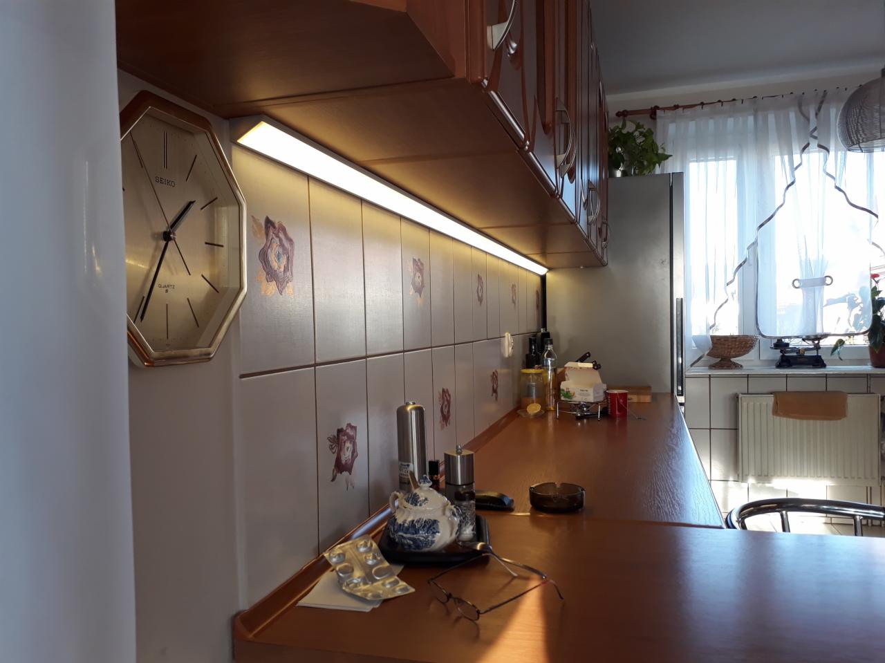 Kuchnia z oświetleniem LED podszafkowym, widoczny zegar Seiko na ścianie z kwiatowym wzorem, drewniane szafki i blat, okno z firanką w tle.