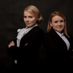 Portret dwóch kobiet w czarnych marynarkach i białych koszulach, stojących plecami do siebie na ciemnym tle, z założonymi rękami i subtelnym uśmiechem.