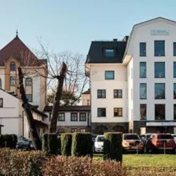 Widok zewnętrzny budynku kancelarii prawnej z zaparkowanymi samochodami, otoczonego zielenią i innymi budynkami.