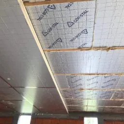 Izolacja termiczna poddasza z folią aluminiową i widocznymi krokwiami, z logo producenta GOR-STAL na folii, w trakcie montażu, widok od dołu na sufit poddasza.