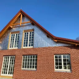 Dom w trakcie budowy z widoczną czerwoną cegłą, oknami z białymi ramami i folią izolacyjną na poddaszu, drewnianą konstrukcją dachu oraz częściowo zamontowaną rynną.