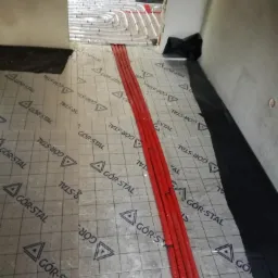 Montaż ogrzewania podłogowego: czerwone rury ułożone na izolacji z nadrukowanym logo GÓR-STAL, widok na rozdzielacz w sąsiednim pomieszczeniu, czarna folia wzdłuż ściany.