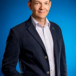 Portret uśmiechniętego mężczyzny w granatowej marynarce i jasnej koszuli na jednolitym niebieskim tle.