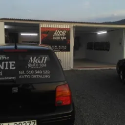 Samochód z naklejką reklamującą pranie tapicerki samochodowej i meblowej na tle myjni samochodowej z napisem 'AUTO DETAILING' na ścianie.