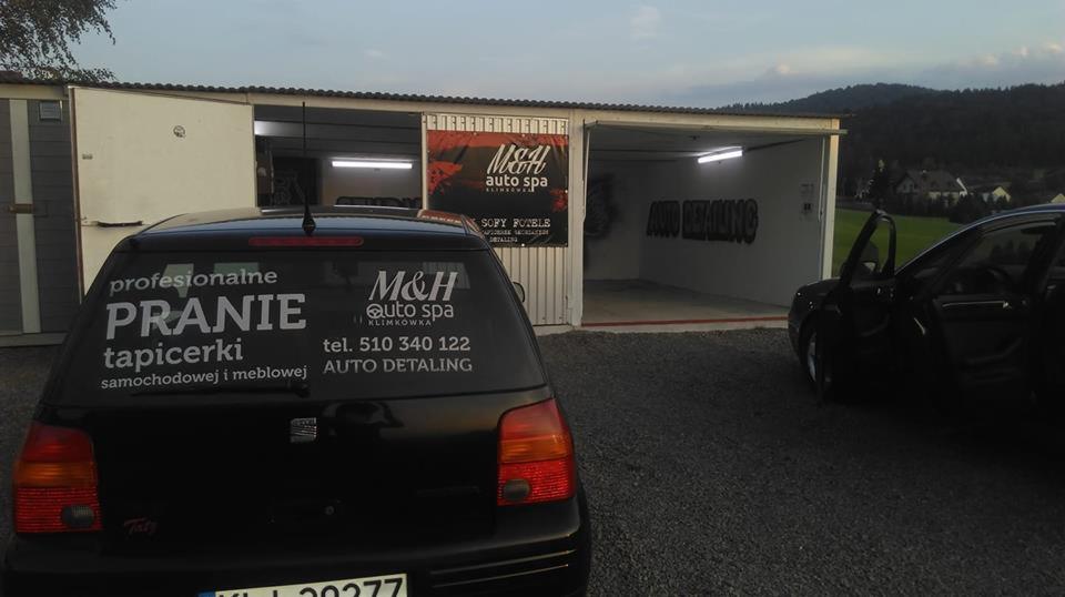 Samochód z naklejką reklamującą pranie tapicerki samochodowej i meblowej na tle myjni samochodowej z napisem 'AUTO DETAILING' na ścianie.
