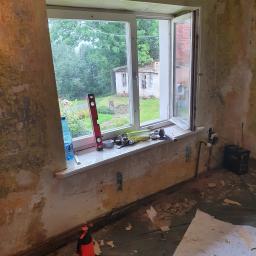 Tomasz Król Usługi Remontowo-Budowlane - Pokój w trakcie remontu: odsłonięte ściany, otwarte okno z widokiem na zieleń, narzędzia na parapecie, folia ochronna na podłodze pokryta resztkami