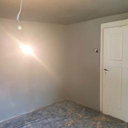 Tomasz Król Usługi Remontowo-Budowlane - Pokój w trakcie remontu, ściany pomalowane na szaro, podłoga zabezpieczona folią malarską, widoczne wystające kable z żarówką, białe drzwi z czarną klamką.