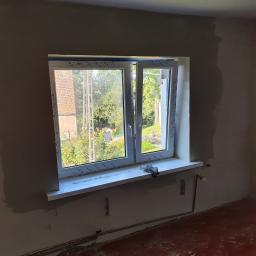 Tomasz Król Usługi Remontowo-Budowlane - Wnętrze pokoju w trakcie remontu: świeżo osadzone okno PCV z widokiem na ogród, otynkowane ściany w szarym odcieniu, wystające rury instalacji grzewczej, podłoga z czerwonej deski.