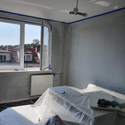 Tomasz Król Usługi Remontowo-Budowlane - Pokój w trakcie przygotowań do malowania: ściany zagruntowane, oklejone taśmą malarską, meble zabezpieczone folią ochronną, widoczne rękawice robocze i nożyk.