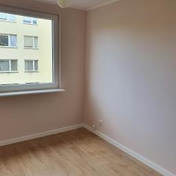 Tomasz Król Usługi Remontowo-Budowlane - Pomalowane na różowo puste pomieszczenie z drewnianą podłogą, widok na budynek z okna, surowa żarówka zwisająca z sufitu.