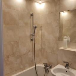 Tomasz Król Usługi Remontowo-Budowlane - Wykończona łazienka z beżowymi płytkami imitującymi kamień, białą wanną i umywalką na postumencie, oświetlona punktowymi lampami sufitowymi.