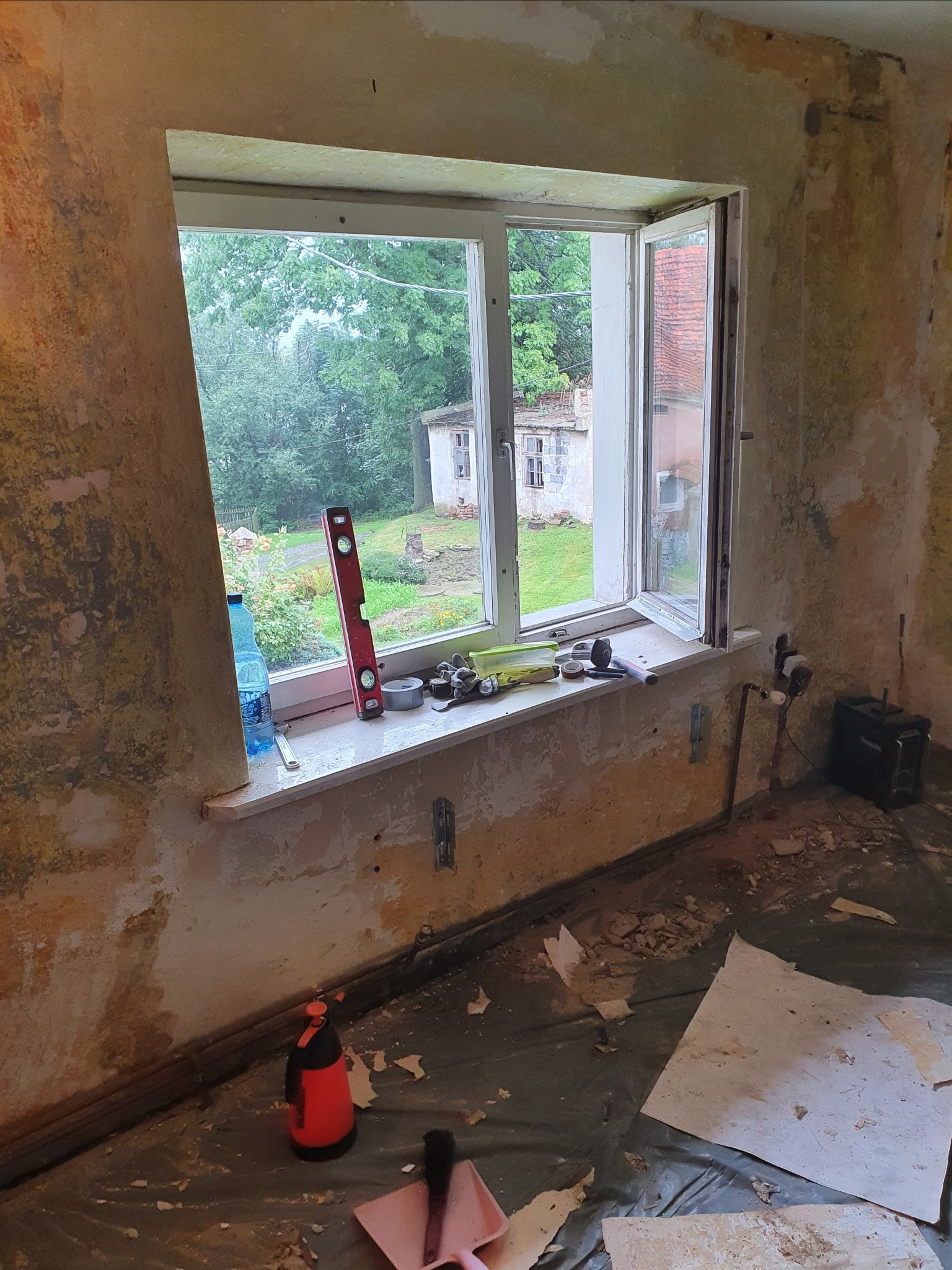 Pokój w trakcie remontu: odsłonięte ściany, otwarte okno z widokiem na zieleń, narzędzia na parapecie, folia ochronna na podłodze pokryta resztkami