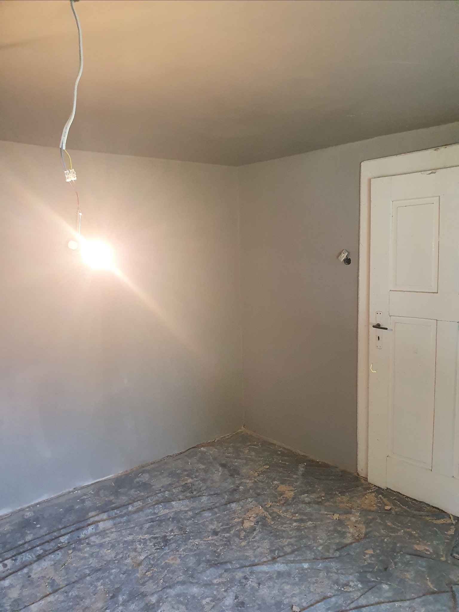 Pokój w trakcie remontu, ściany pomalowane na szaro, podłoga zabezpieczona folią malarską, widoczne wystające kable z żarówką, białe drzwi z czarną klamką.