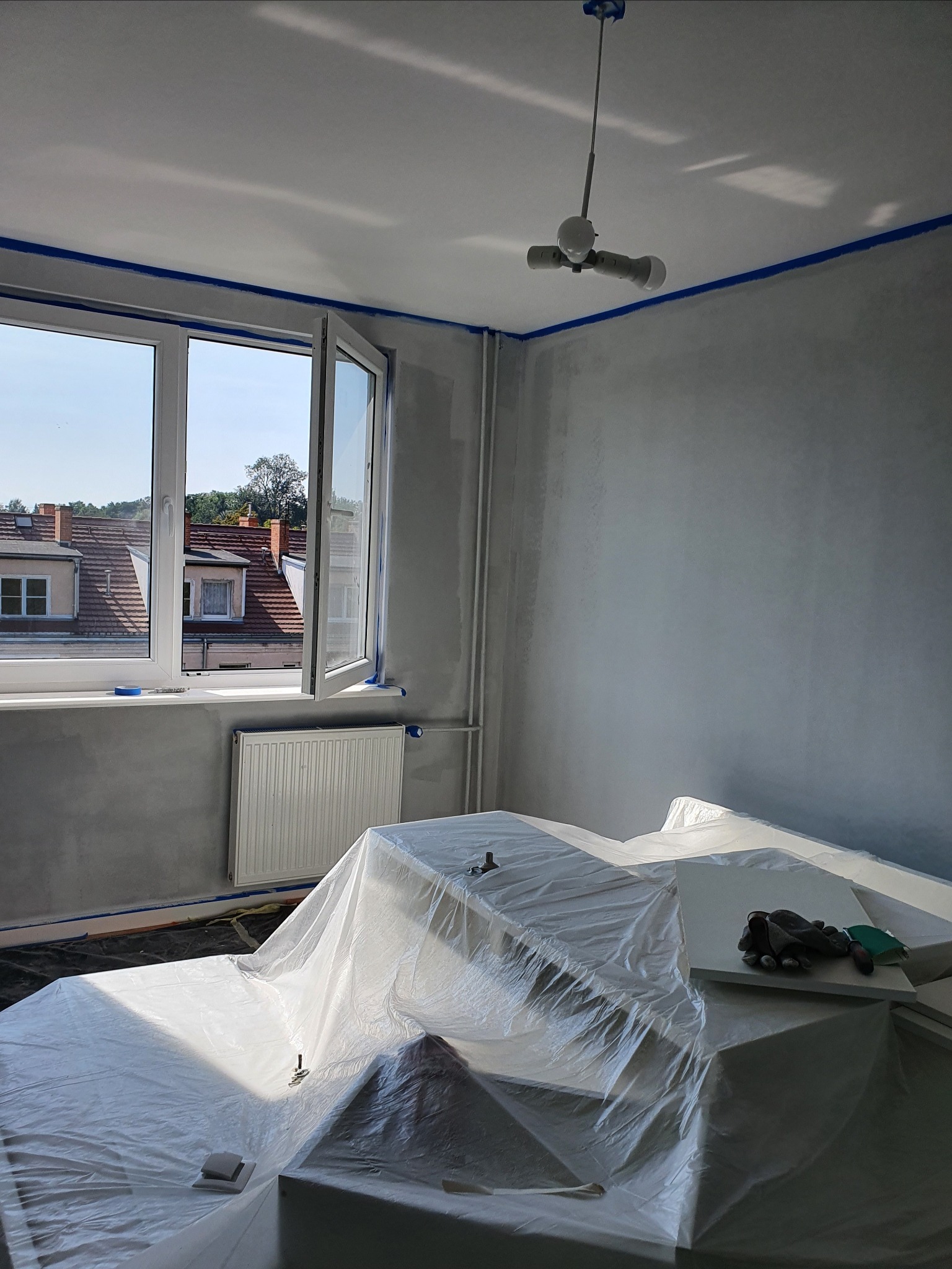 Pokój w trakcie przygotowań do malowania: ściany zagruntowane, oklejone taśmą malarską, meble zabezpieczone folią ochronną, widoczne rękawice robocze i nożyk.