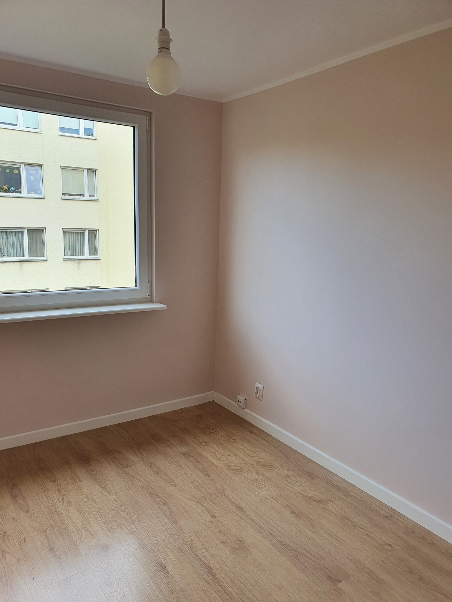Pomalowane na różowo puste pomieszczenie z drewnianą podłogą, widok na budynek z okna, surowa żarówka zwisająca z sufitu.