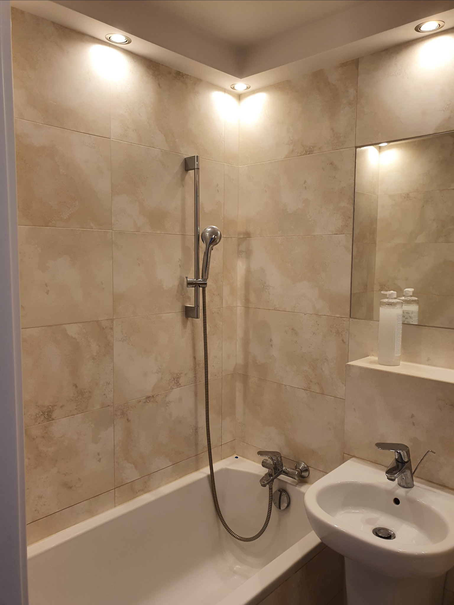 Wykończona łazienka z beżowymi płytkami imitującymi kamień, białą wanną i umywalką na postumencie, oświetlona punktowymi lampami sufitowymi.