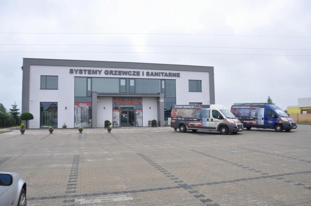 Budynek firmy z napisem 'Systemy grzewcze i sanitarne' oraz dwa samochody dostawcze z logo firmy 'Systemy Grzewcze Karles' na parkingu z kostki brukowej.