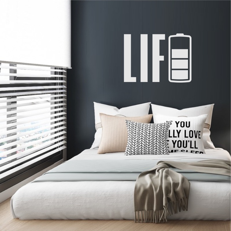 Nowoczesna sypialnia z białym łóżkiem, dekoracyjnymi poduszkami i napisem 'LIFE' z symbolem baterii na ciemnej ścianie, widok z okna przez żaluzje.
