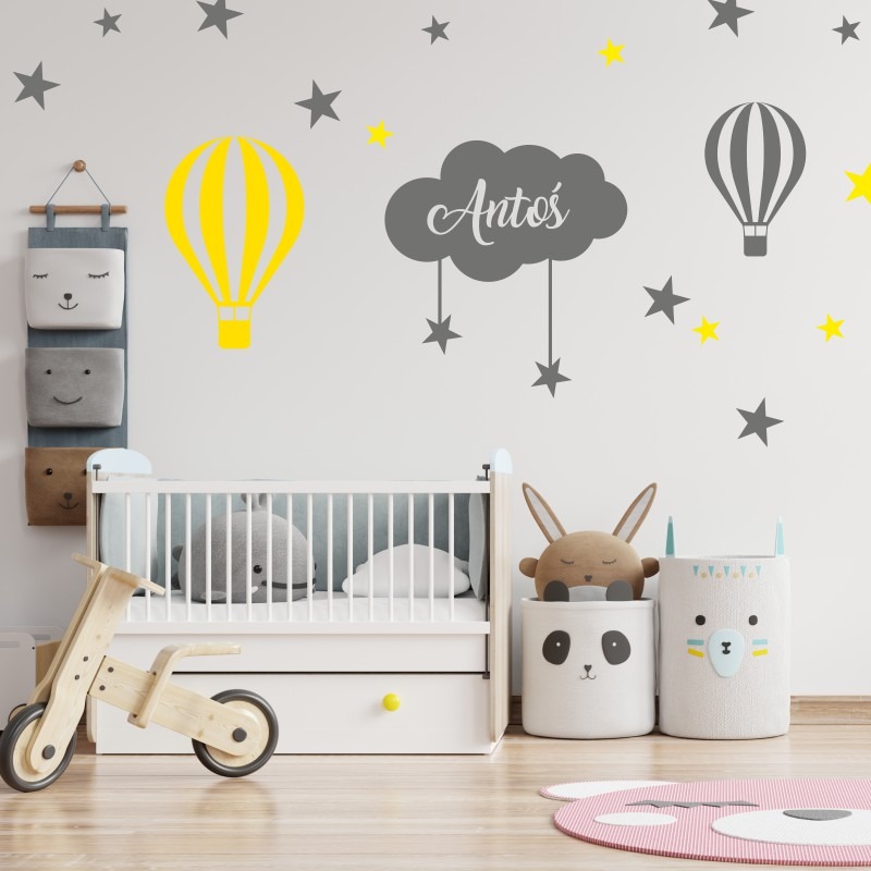 Dziecięcy pokój z białym łóżeczkiem, drewnianym rowerkiem, pojemnikami na zabawki w kształcie zwierząt i naklejkami na ścianie z motywem chmurki z imieniem 'Antoś', balonów i gwiazd.