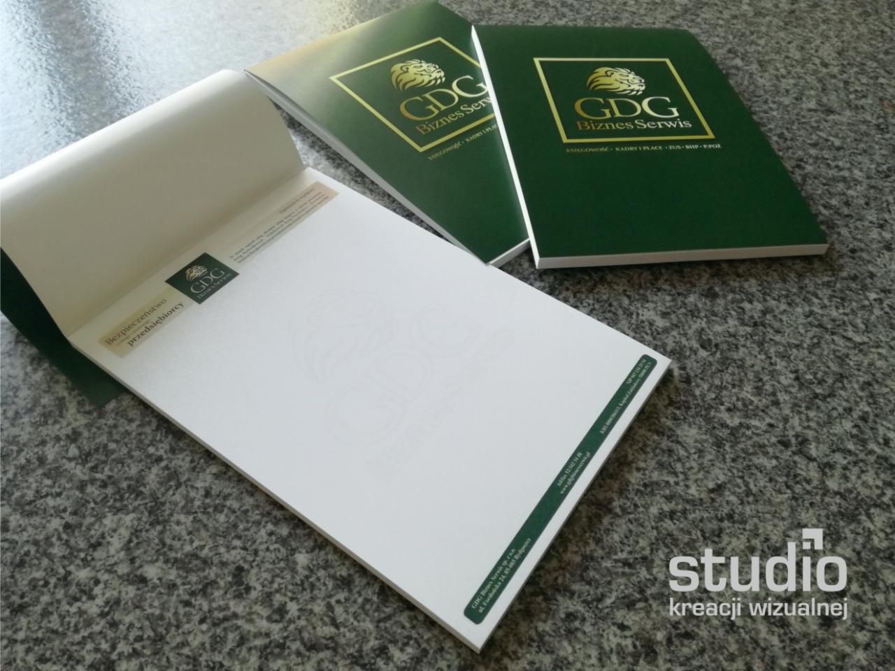 Zestaw firmowych notatników GGD Biznes Serwis w kolorze ciemnej zieleni ze złotym logo, jeden otwarty z widocznym nadrukiem 'Bezpieczeństwo przedsiębiorcy'.