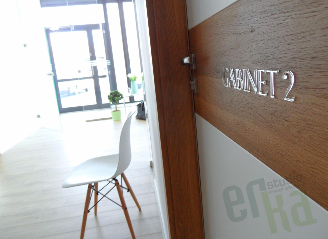 Oznakowanie drewnianych drzwi z napisem 'GABINET 2' wykonanym z przezroczystego akrylu, widok z korytarza z krzesłem i rośliną doniczkową w tle.