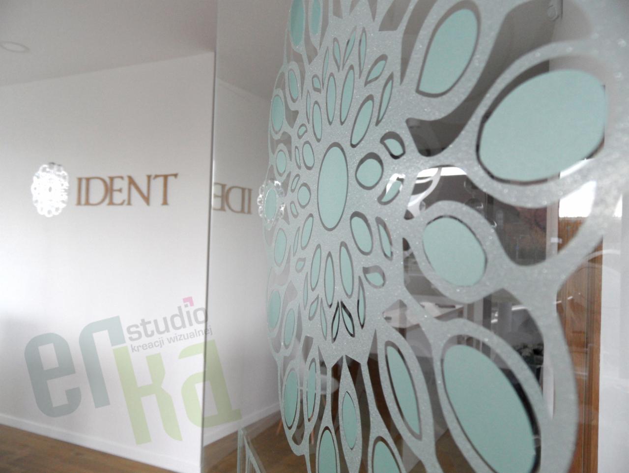 Szklane drzwi z ozdobnym, matowym wzorem kwiatowym w biurze, widoczne logo firmy na ścianie w tle.