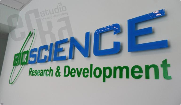 Ścienne litery przestrzenne z pleksi, w kolorach zielonym i niebieskim, tworzące nazwę firmy BIOSCIENCE Research & Development, z efektem połysku i taśmą zabezpieczającą na górnej krawędzi litery E.