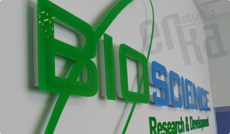 Zielono-niebieski, trójwymiarowy szyld z nazwą firmy 'BioScience Research & Development' zamontowany na białej ścianie, z widocznym logo 'Studio Kreacji Wizualnej' w tle.