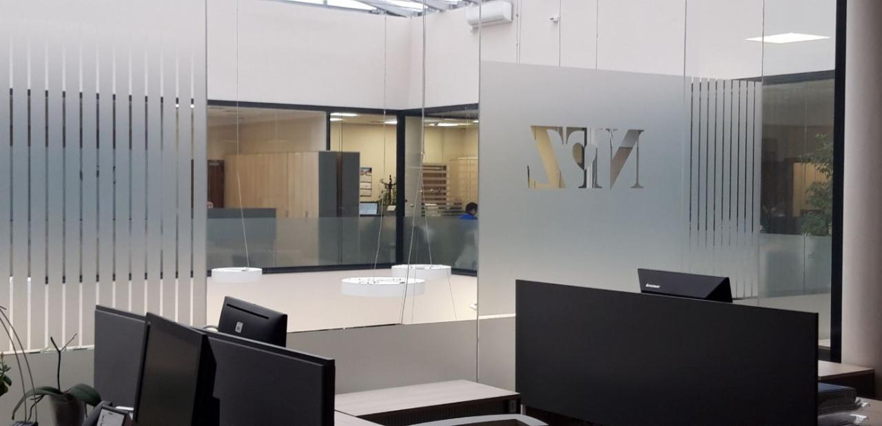 Nowoczesne biuro z przeszklonymi ścianami i logo firmy, widoczne stanowiska pracy z monitorami, oświetlenie sufitowe w kształcie okręgów.