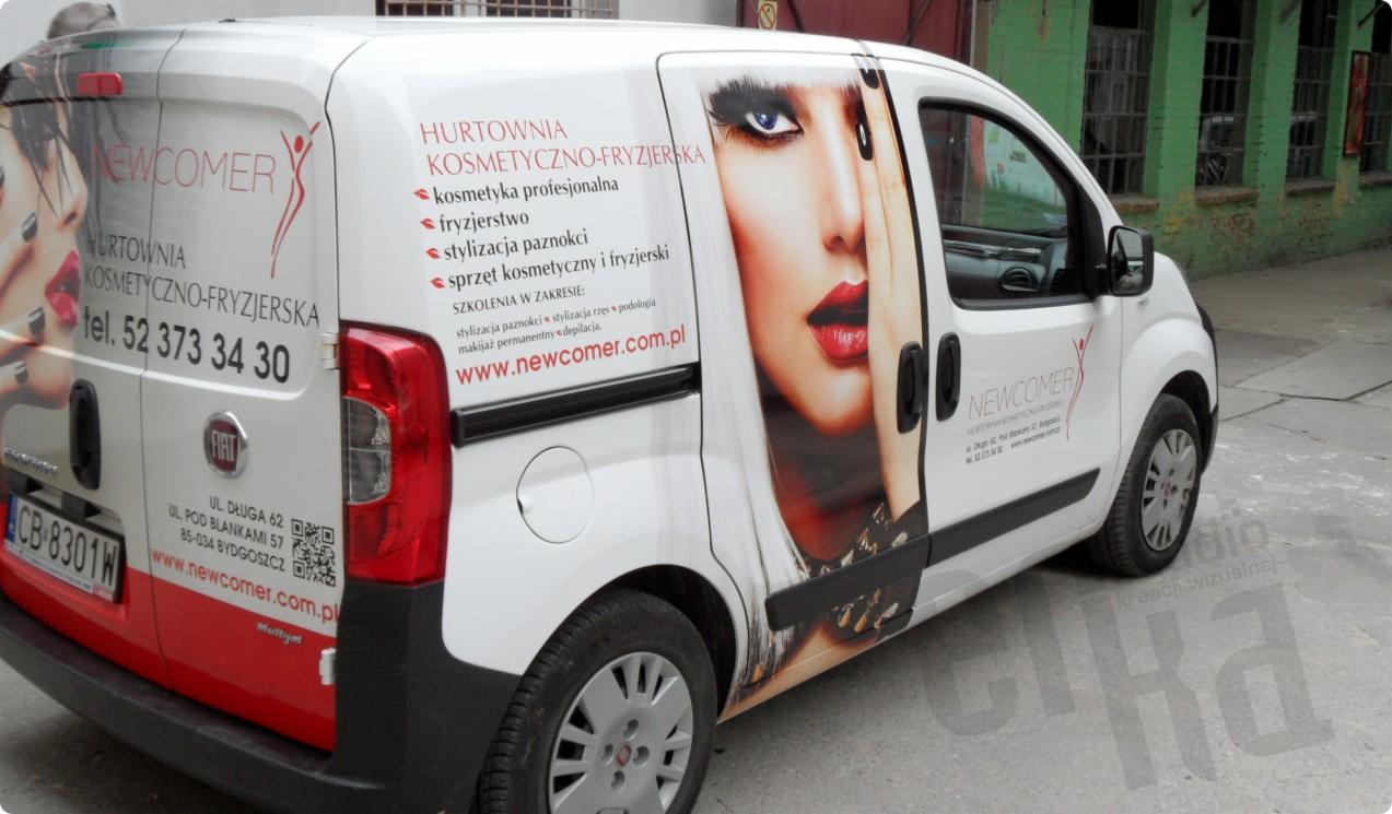 Oklejony samochód firmowy z reklamą hurtowni kosmetyczno-fryzjerskiej Newcomer, prezentujący grafikę twarzy kobiety z mocnym makijażem i informacjami o ofercie, zaparkowany na tle zielonego budynku.