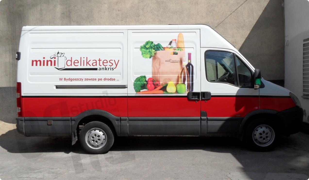 Biało-czerwony samochód dostawczy z grafiką warzyw, owoców i butelki wina oraz logo 'mini delikatesy ankris' na białej części karoserii.