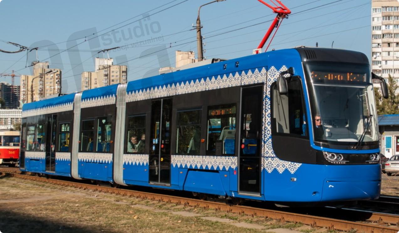 Niebieski tramwaj z ludowym wzorem na tle miejskiego krajobrazu, widoczny motorniczy za szybą.