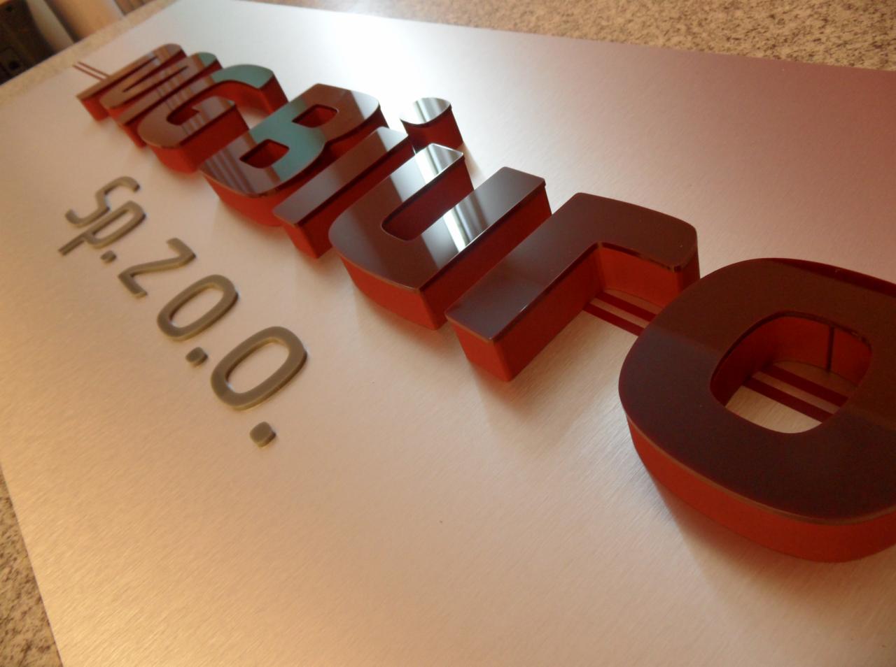 Trójwymiarowe, błyszczące litery logo firmy w odcieniach czerwieni i brązu na metalowej płycie, obok napis 'sp. z o.o.' w szarym kolorze.